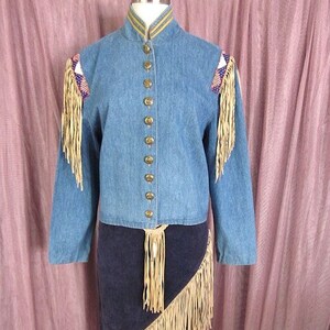 May include: A denim jacket with fringe detailing on the shoulders and a matching denim skirt with fringe trim. The jacket has a stand-up collar and a button-up front.