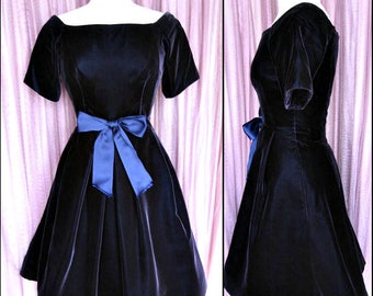 May include: A navy blue velvet dress with a bateau neckline and short sleeves. The dress features a blue satin bow at the waist. The skirt is flared, creating a classic silhouette. The dress is displayed on a mannequin.