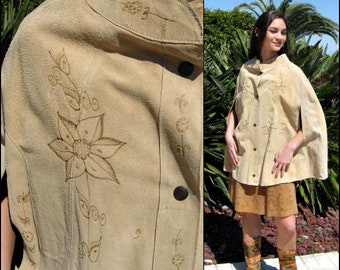 May include: A tan suede cape with floral embroidery. The cape has a collar and a button closure. The model is wearing the cape over a brown skirt and brown boots.