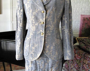 May include: A gray and gold patterned blazer and skirt suit. The blazer has a single-breasted closure with gold buttons. The skirt is knee-length and has a straight silhouette.