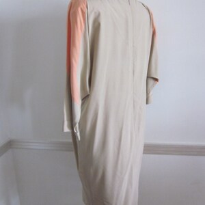 May include: A beige dress with a peach colored panel on the sleeves. The dress has a simple, elegant design and a straight silhouette.