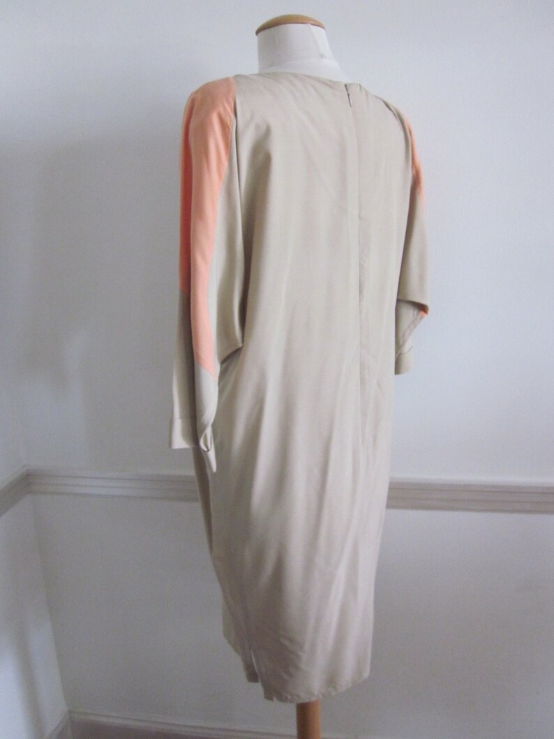 May include: A beige dress with a peach colored panel on the sleeves. The dress has a simple, elegant design and a straight silhouette.