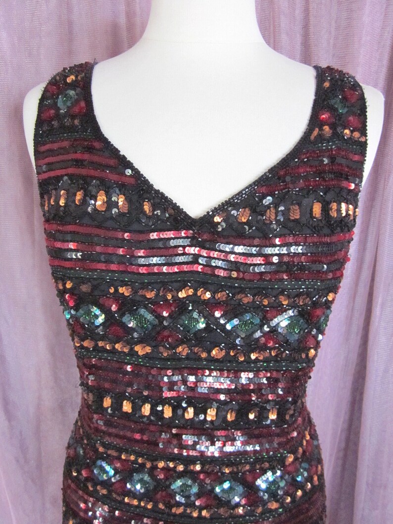 Puede incluir: Un vestido sin mangas de lentejuelas con escote en V. El vestido es negro con lentejuelas rojas, doradas y verdes dispuestas en un patr&oacute;n geom&eacute;trico.