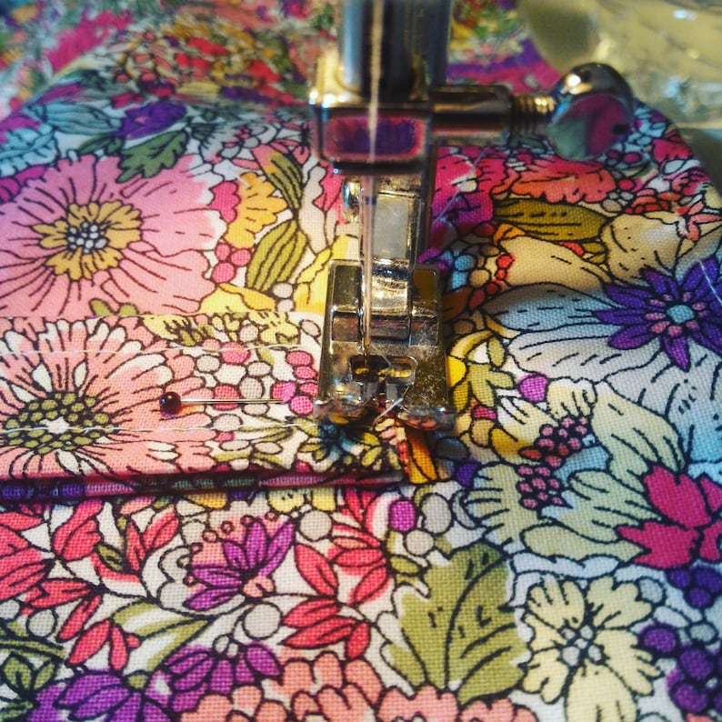 May include: Close-up of a sewing machine needle going through a floral fabric with a pink flower pattern. The fabric is being sewn with a white thread.