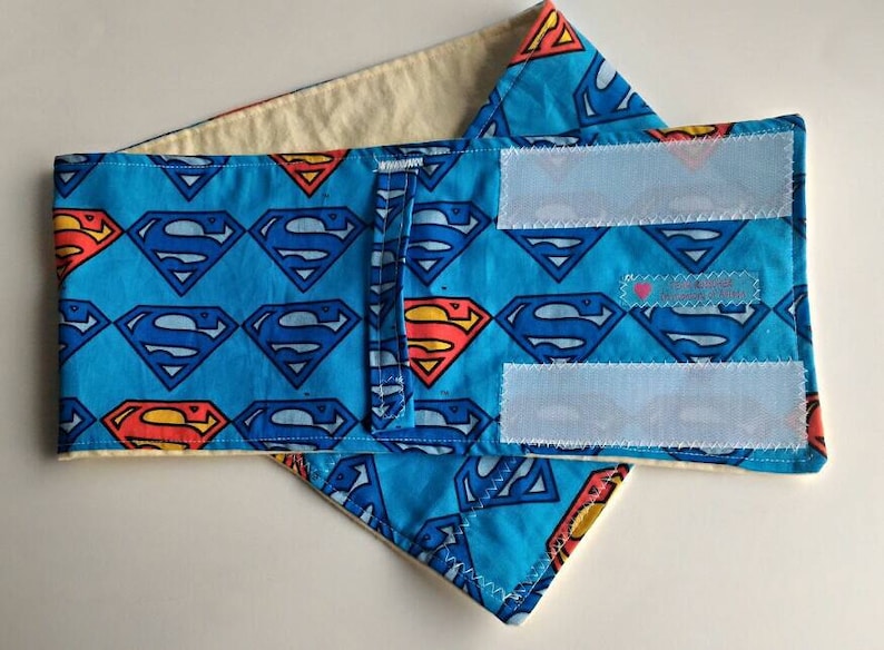 May include: A blue bandana with a white background and a repeating pattern of Superman logos. The bandana has two white velcro straps on the back.