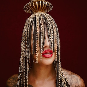 Può includere: Una donna indossa un copricapo dorato con una frangia di catene che le copre il viso. Il copricapo ha una parte superiore rotonda ed è decorato con piccoli strass trasparenti. La donna indossa il rossetto rosso e ha un tatuaggio sulla spalla.