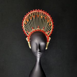 Puede incluir: Un tocado dorado con detalles rojos y un diseño en forma de abanico. El tocado presenta joyas y cuentas rojas, con un patrón decorativo alrededor de la base. El tocado se exhibe sobre una cabeza de maniquí negra sobre un fondo negro.