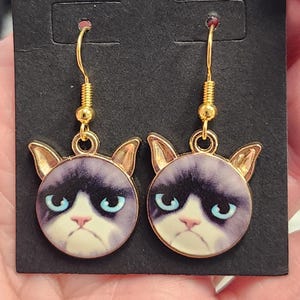 May include: A pair of gold dangle earrings featuring a grumpy cat face with blue eyes. The cat's face is printed on a round white background with a gold rim.