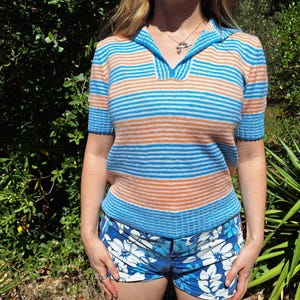 May include: A vintage-style knit top with a polo collar, featuring horizontal stripes in shades of blue and orange. The top is paired with blue floral print shorts. The outfit is suitable for casual wear.