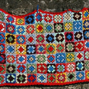 Puede incluir: Una manta de ganchillo colorida con un borde rojo, colgada de un tendedero contra una pared de piedra.