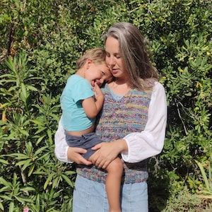 Peut inclure: Une femme portant une chemise blanche avec un gilet en tricot coloré tient un jeune enfant dans ses bras. L'enfant porte une chemise bleue et un short bleu foncé. La femme et l'enfant sont debout devant un fond vert feuillu.