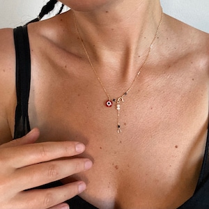 May include: A gold chain necklace with a red and white evil eye charm, a pearl, and black beads. The necklace is layered with a smaller gold chain with a circle charm.