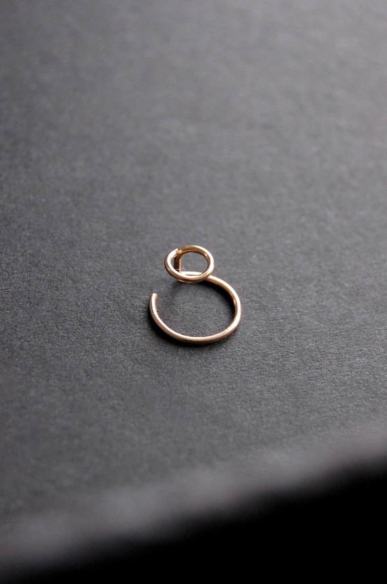May include: A gold-colored, minimalist earring with a unique design. The earring features a small, circular top connected to a larger, open-ended hoop. The jewelry is set against a dark background.