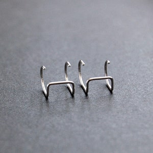 May include: Three silver hoop earrings with a curved, open design. The earrings are arranged in a row on a gray surface.