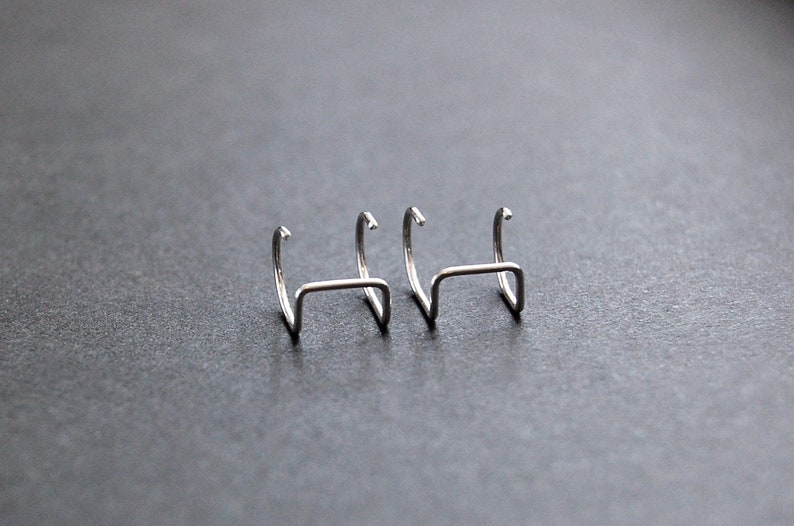 May include: Three silver hoop earrings with a curved, open design. The earrings are arranged in a row on a gray surface.