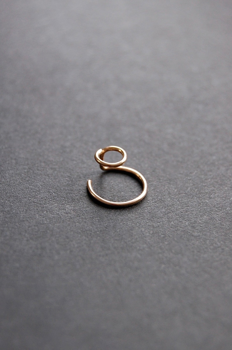 May include: A delicate gold-colored ear cuff. The ear cuff features a small, open circle at the top, connected to a larger, open circle that curves around. The jewelry is set against a dark gray background.