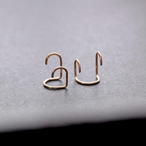 May include: Two small, gold-toned metal ear cuffs. One cuff is shaped like a simple heart outline, while the other is a plain, open-ended U-shaped band. Both pieces feature a smooth, polished finish and are designed to sit securely on the ear cartilage.