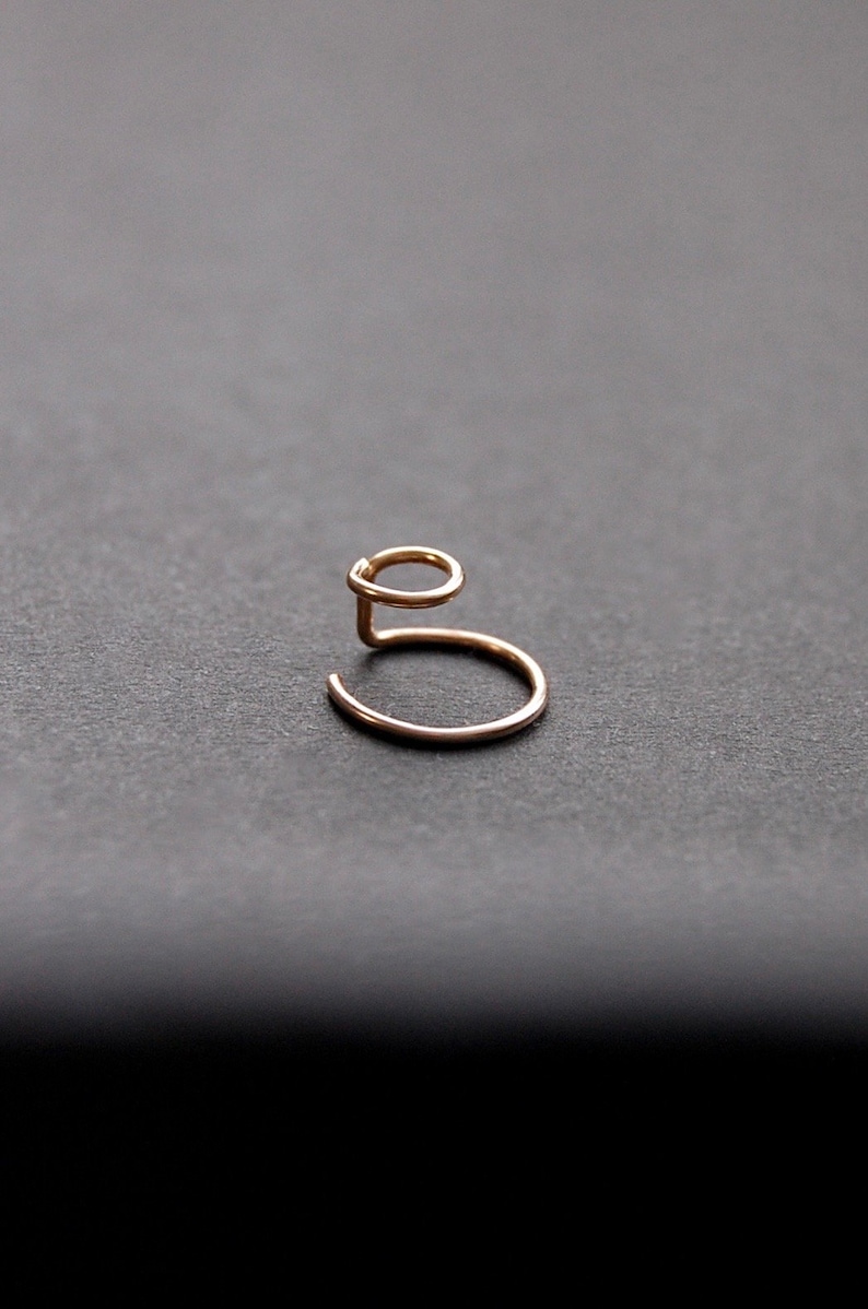 May include: A delicate, gold-colored ear cuff. The ear cuff has a unique, minimalist design with a curved base and a loop that curves upwards. The jewelry is set against a dark gray background.