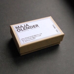 K&ouml;nnte beinhalten: Eine kleine braune Schachtel aus Pappe mit einem wei&szlig;en Etikett, auf dem "MAJA OLENDER handgemachter Schmuck" steht und Kontaktinformationen enthalten sind. Das Etikett tr&auml;gt au&szlig;erdem den Schriftzug "DANKE!"