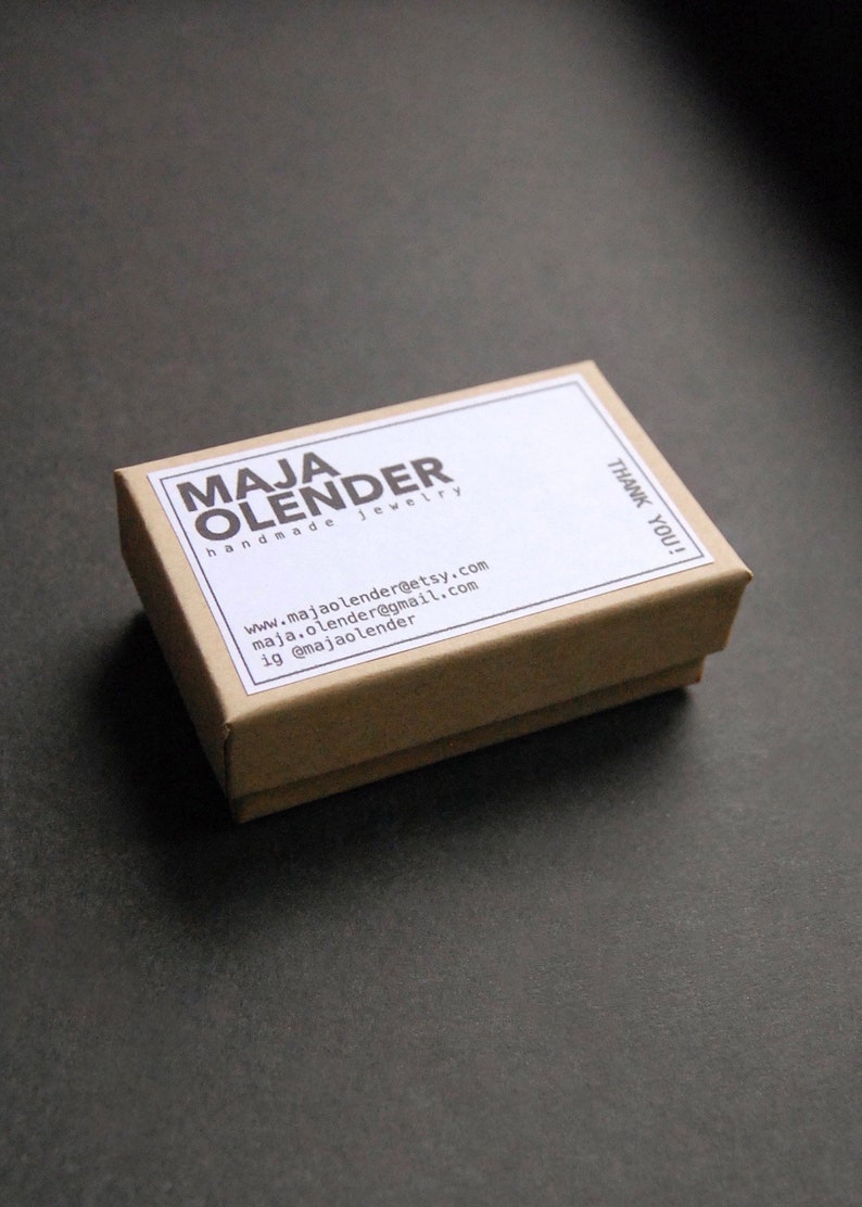 K&ouml;nnte beinhalten: Eine kleine braune Schachtel aus Pappe mit einem wei&szlig;en Etikett, auf dem "MAJA OLENDER handgemachter Schmuck" steht und Kontaktinformationen enthalten sind. Das Etikett tr&auml;gt au&szlig;erdem den Schriftzug "DANKE!"