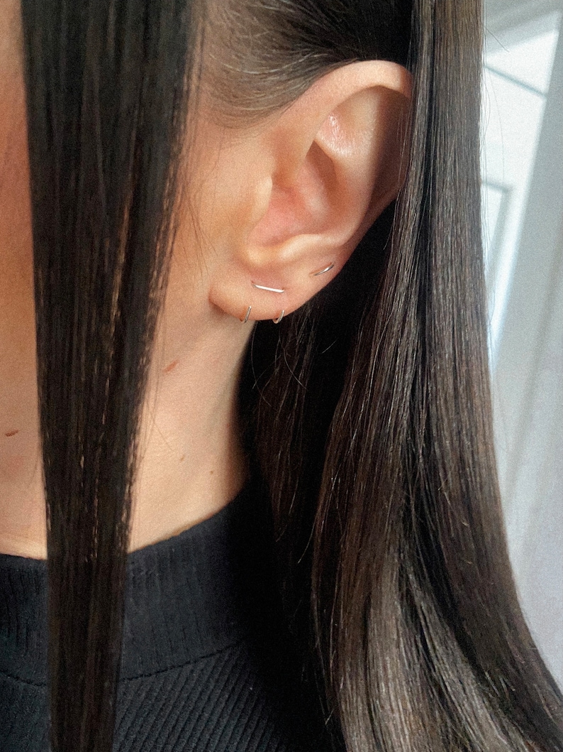 May include: A close-up of a person's ear with three small silver hoop earrings. The earrings are simple and delicate, and they are worn in the upper lobe of the ear.