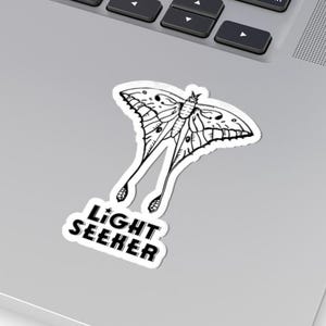 May include: A white sticker featuring a detailed black and white illustration of a moth with long, flowing tails. The sticker has the text "LIGHT SEEKER" in a bold, sans-serif font below the moth design. The sticker is placed on a silver laptop.