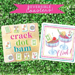 May include: Two square coasters with a gold and white background. One coaster features a colorful mahjong tile design with the words "crack dot bam". The other coaster features a pink and green border with a white background and a drawing of two cocktails with the words "clink!"