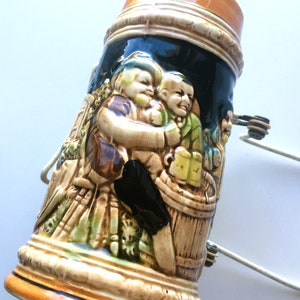 May include: A ceramic beer stein with a detailed relief of figures in a tavern setting. The stein features a brown, black, and tan color scheme with a handle and a decorative rim. The relief depicts people, barrels, and mugs.