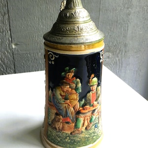 May include: A vintage beer stein with a detailed relief scene of people. The ceramic stein has a dark blue background with a silver metal lid and handle. The scene depicts people in traditional clothing, with a dog, and musical instruments.