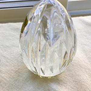 May include: A clear, egg-shaped glass paperweight featuring a faceted, vertical cut pattern across its surface. A small, gold-colored rectangular sticker is adhered to the upper left side of the glass object, displaying faint black text.