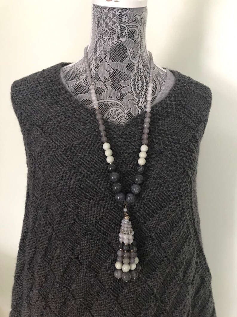 May include: A gray, white, and black beaded necklace with a tassel pendant. The necklace is on a gray sweater.