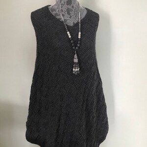 May include: A gray sleeveless sweater with a cable knit pattern. The sweater is on a white mannequin and has a long, flowing shape. A gray and white beaded necklace with a tassel hangs from the sweater.