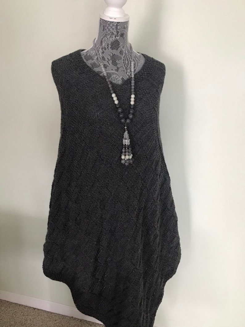 May include: A gray sleeveless sweater with a cable knit pattern. The sweater is on a white mannequin and has a long, flowing shape. A gray and white beaded necklace with a tassel hangs from the sweater.