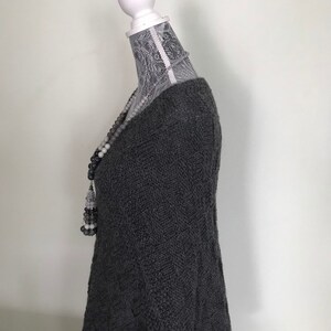 May include: A gray knitted sweater with a loose fit and a v-neck. The sweater is on a white mannequin with a lace covering the head and neck.