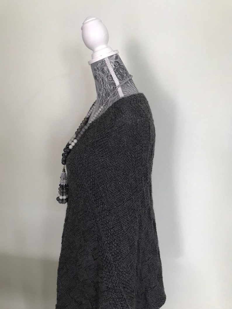 May include: A gray knitted sweater with a loose fit and a v-neck. The sweater is on a white mannequin with a lace covering the head and neck.