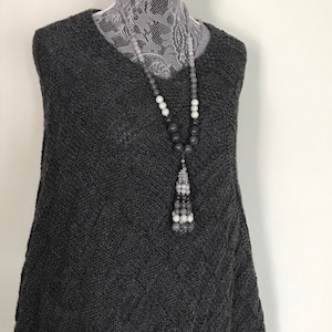 May include: A gray knitted sleeveless sweater with a long beaded necklace featuring a tassel. The necklace has gray, white, and black beads.