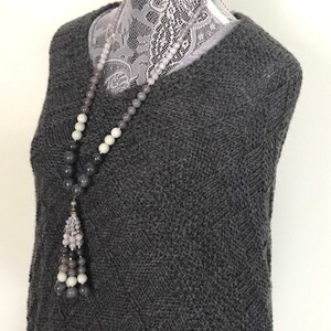 May include: A gray knitted sweater with a long gray, white, and black beaded necklace with a tassel.