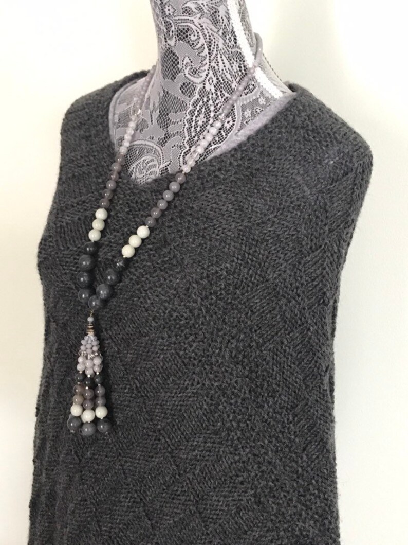 May include: A gray knitted sweater with a long gray, white, and black beaded necklace with a tassel.