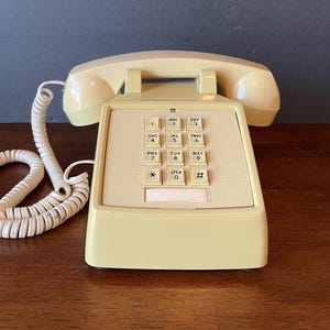 May include: A beige rotary dial phone with a white cord. The phone has a keypad with letters and numbers. The phone is on a wooden surface.