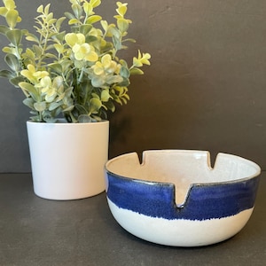 May include: A ceramic ashtray with a white base and a bold blue stripe around the rim. The ashtray has three notches for resting cigarettes. A small white potted plant is in the background.