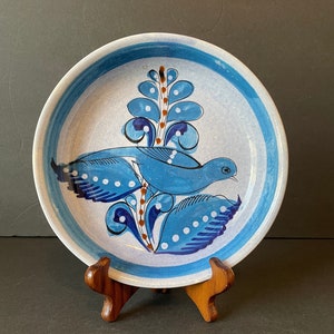 May include: A blue and white ceramic plate with a stylized bird design. The bird is perched on a branch with leaves and flowers. The plate has a white background with blue and brown accents.