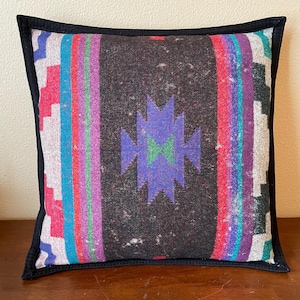 May include: A square throw pillow with a black border and a colorful geometric pattern in shades of pink, blue, green, and purple. The pattern is reminiscent of a traditional Native American design.
