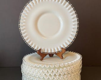 May include: A stack of white, ruffled-edge plates with a clear, decorative trim. One plate is displayed on a wooden stand. The plates have a classic, vintage aesthetic, suitable for serving food or as decorative pieces.