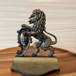 May include: A gold-toned metal lion doorstop with a shield and scroll design. The lion is standing on a base with a curved edge.