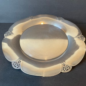 May include: A silver-colored, scalloped-edge plate with a central, raised circular area. The plate's rim features decorative cut-out designs. The plate is likely made of metal and has a reflective surface.