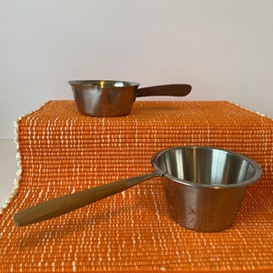 May include: Two small silver pots with wooden handles on an orange and white woven placemat.