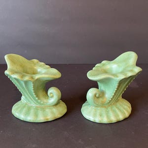 May include: Two matching green ceramic candle holders. Each has a flared top, a curved, scroll-like design, and a textured base. The candle holders are a muted green color and sit on a dark background.