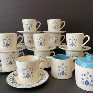 May include: A vintage cream-colored tea set with a blue and green leaf design. The set includes cups, saucers, a creamer, and a sugar bowl with a blue lid. The cups are stacked on top of each other.