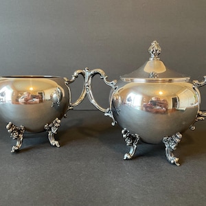May include: A silver creamer and sugar bowl set with ornate handles and feet. The pieces are highly polished and reflect light.