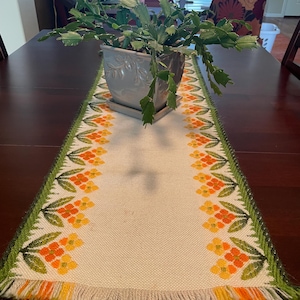 May include: A rectangular table runner featuring a cream-colored woven fabric base with a repeating floral pattern in shades of orange and yellow. The edges are bordered by green foliage designs and finished with a fringed hem in orange, yellow, and green.